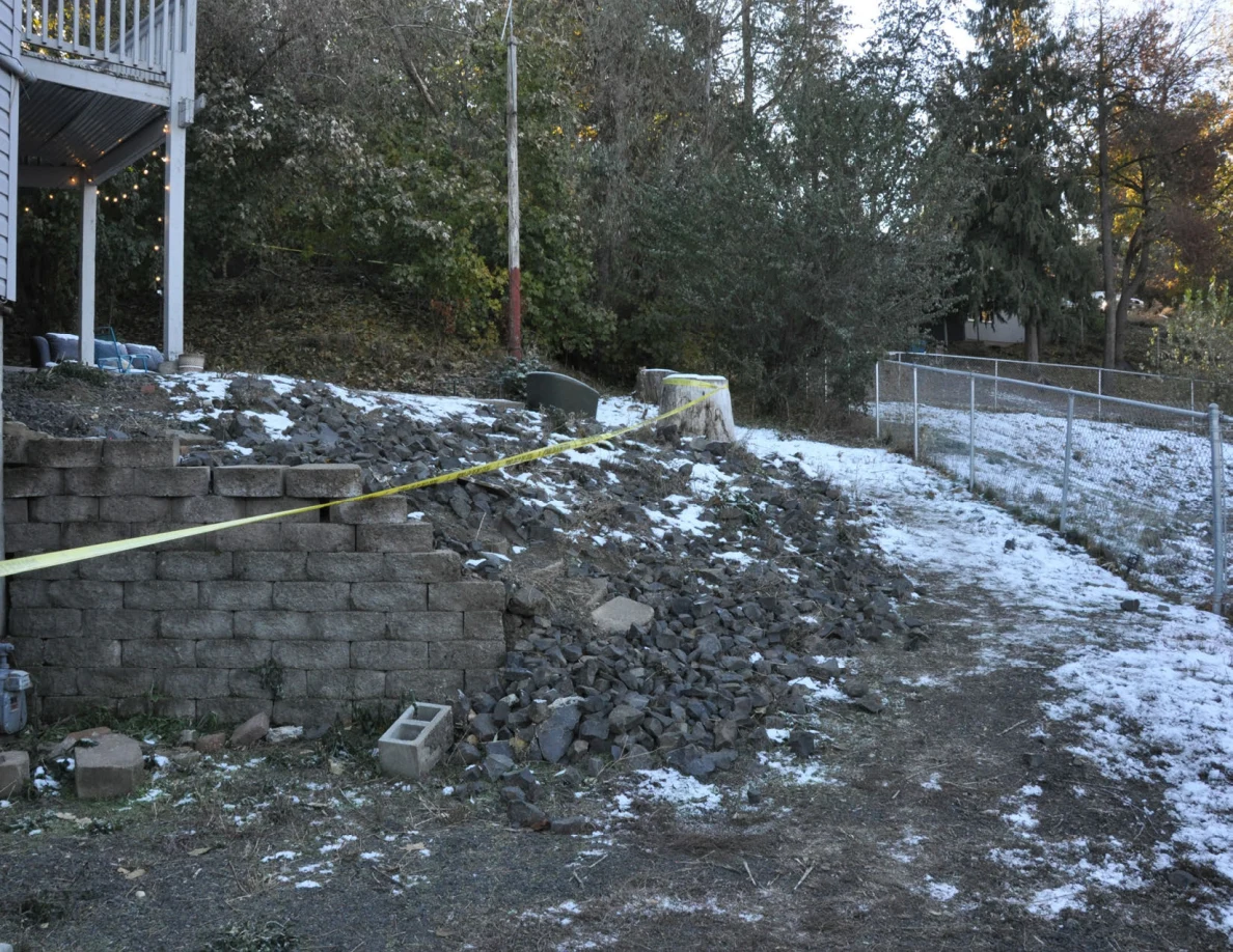 Snow-dusted slope with a retaining wall, yellow tape, chain-link fence and trees.