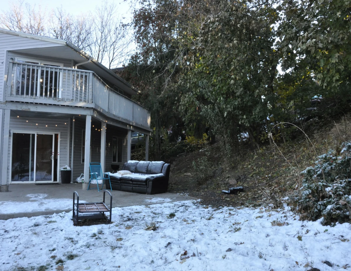 Snowy backyard with a sofa beneath a raised deck, sliding glass doors and an overturned metal frame.