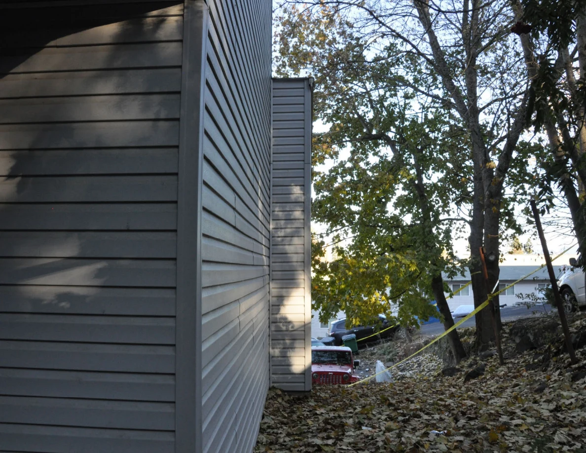 Narrow side yard between house siding and trees, with yellow tape and parked vehicles beyond.