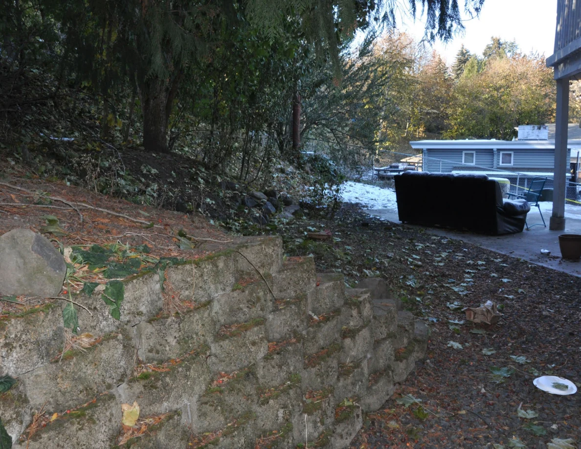Block retaining wall beside a leafy path leading to an outdoor sofa near the house.