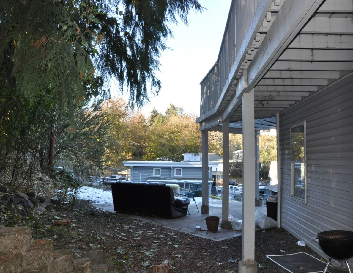 Rear patio beneath a raised deck, with an outdoor sofa, trees and patches of snow.