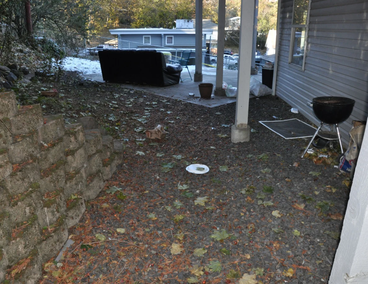 Gravel path beside a retaining wall and house, with a kettle grill, window screen and white plate.