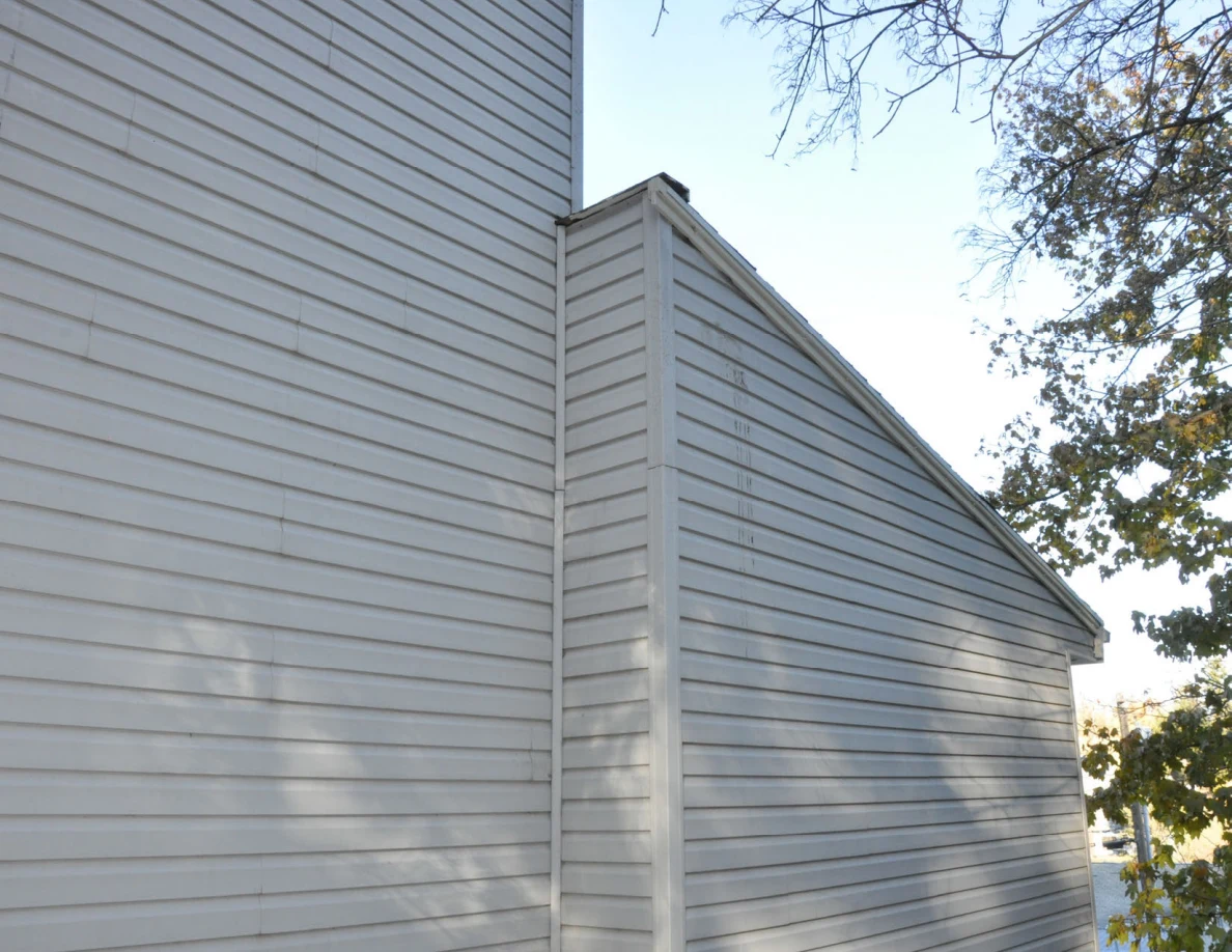 Corner where two gray-sided sections of a house meet beneath roof eaves.