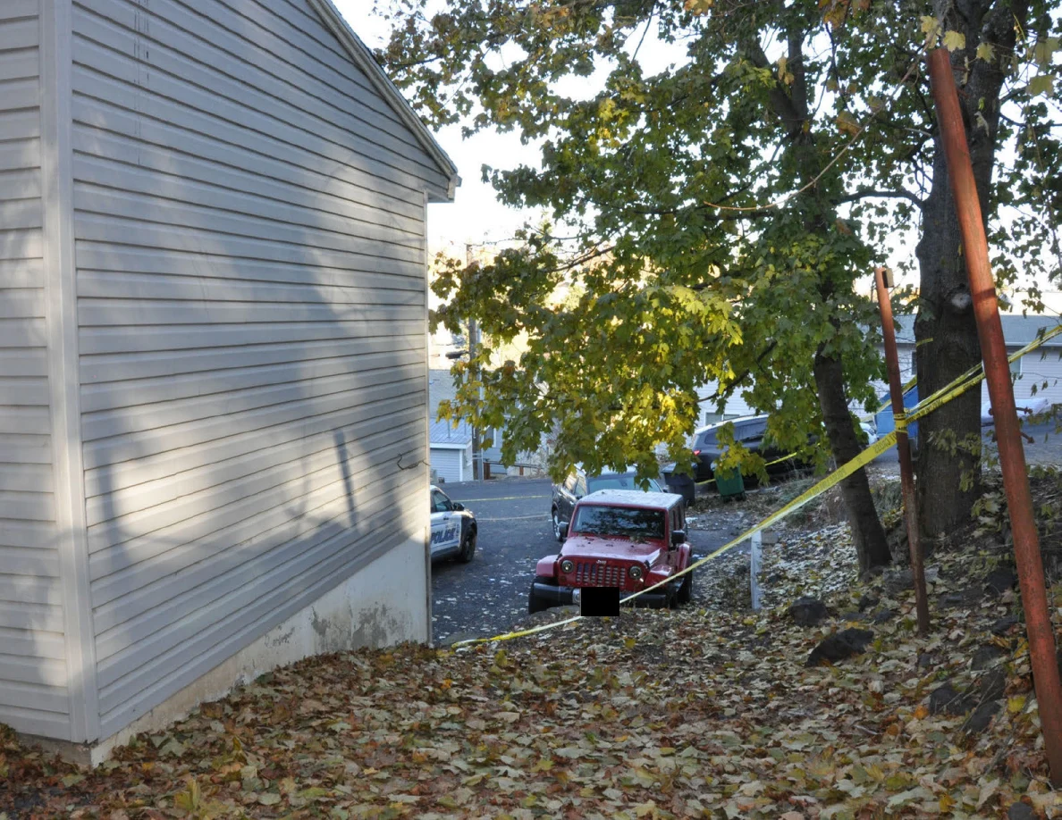 View along house siding toward a red Jeep below a leafy slope crossed by yellow tape.