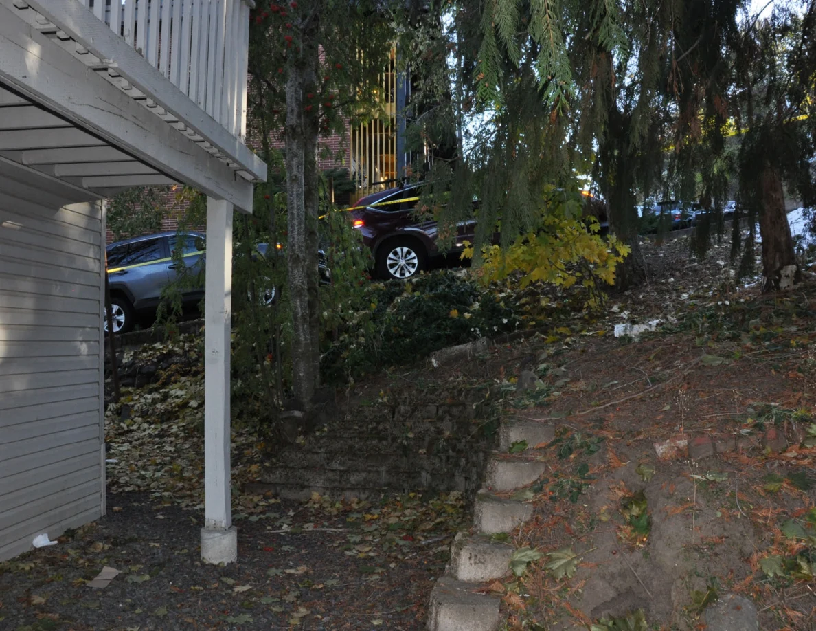 Deck support post beside a retaining wall and wooded slope with parked vehicles beyond.