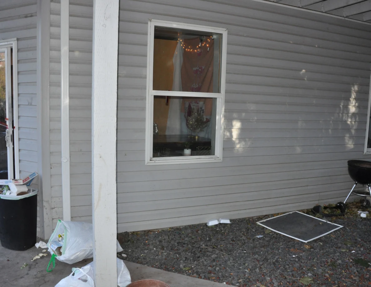 Exterior window in gray siding beneath a deck, with a detached window screen on gravel.