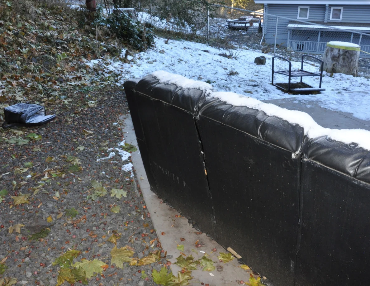 Snow along the back of an outdoor sofa, with an overturned metal frame in the yard.