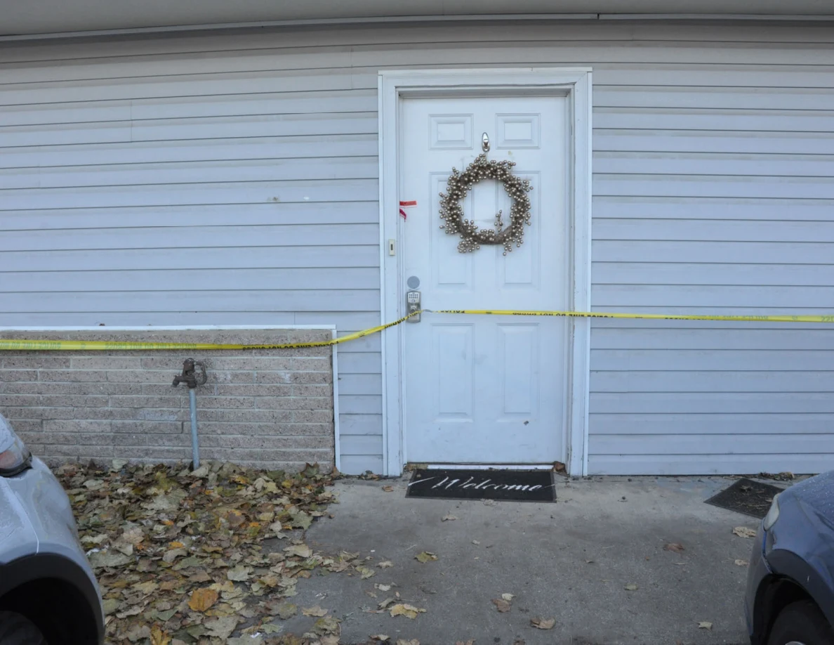 White front door with a wreath and welcome mat, crossed by yellow tape.