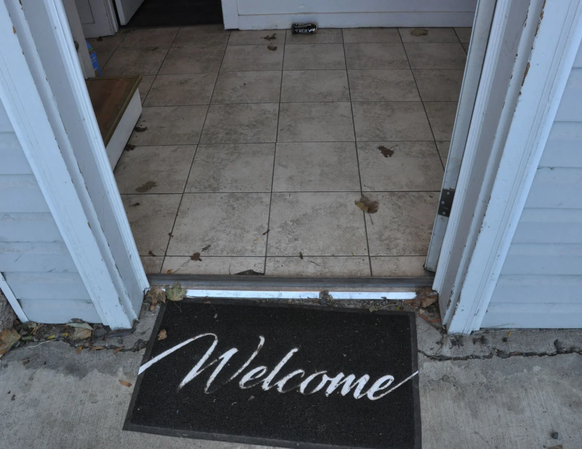 Open front doorway with a black Welcome mat, tiled floor and scattered dry leaves.