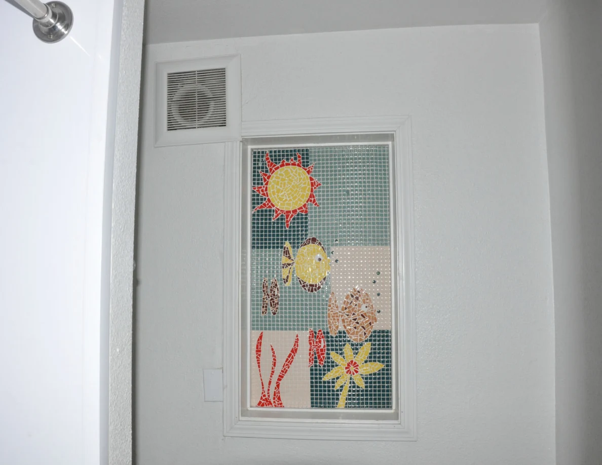 Bathroom wall with a colorful sun-and-fish decorative window panel and ventilation grille.