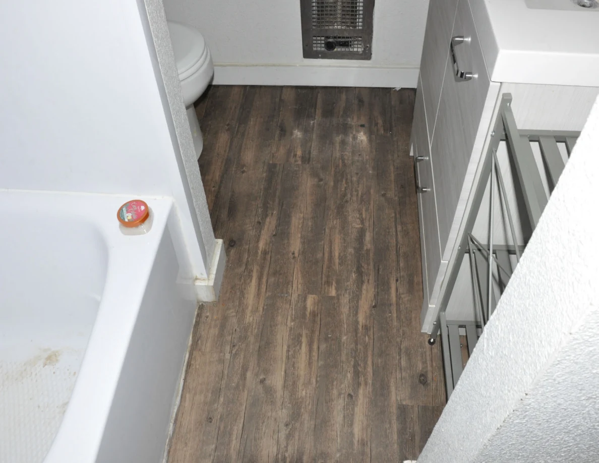 Wood-look bathroom floor between a bathtub and vanity beside a narrow metal shelf.