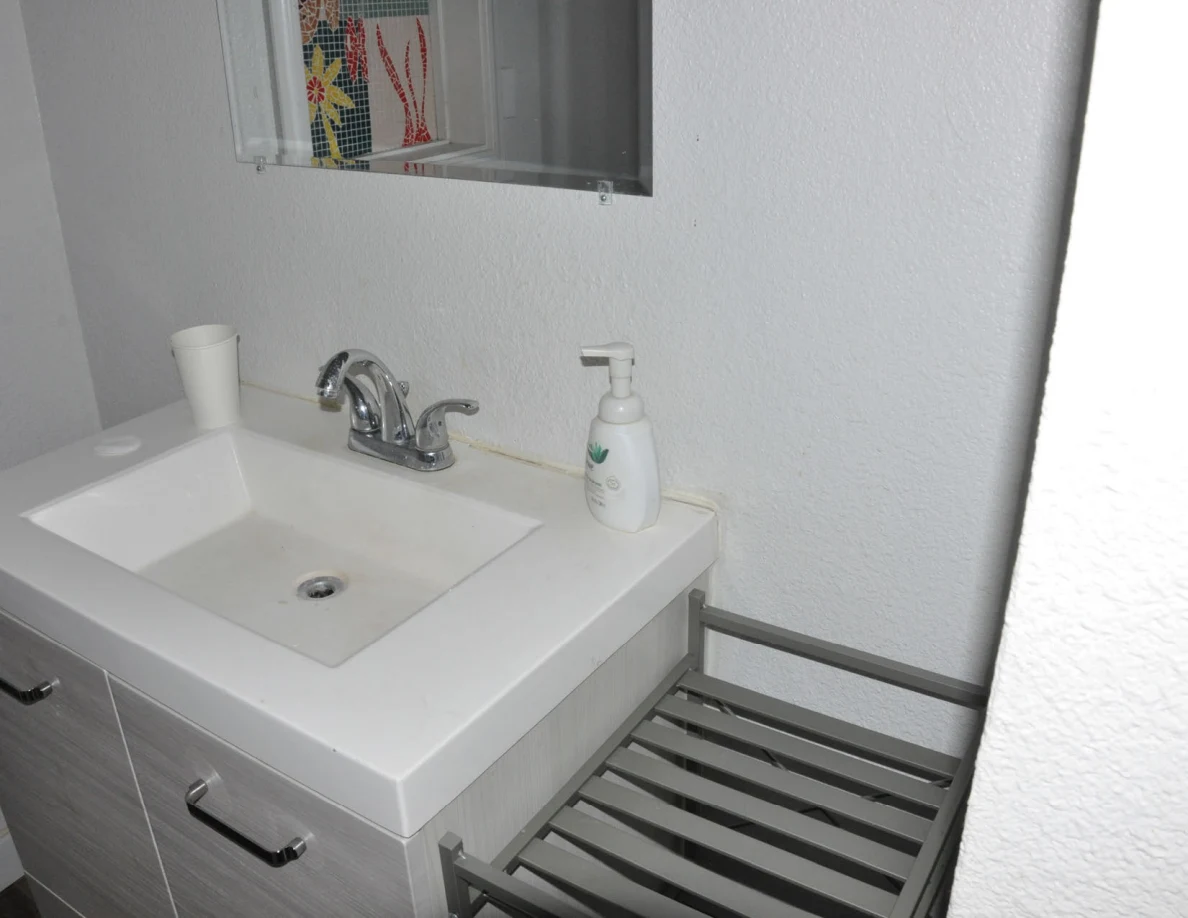 White bathroom sink and chrome faucet with a pump bottle and cup, beside a metal shelf.