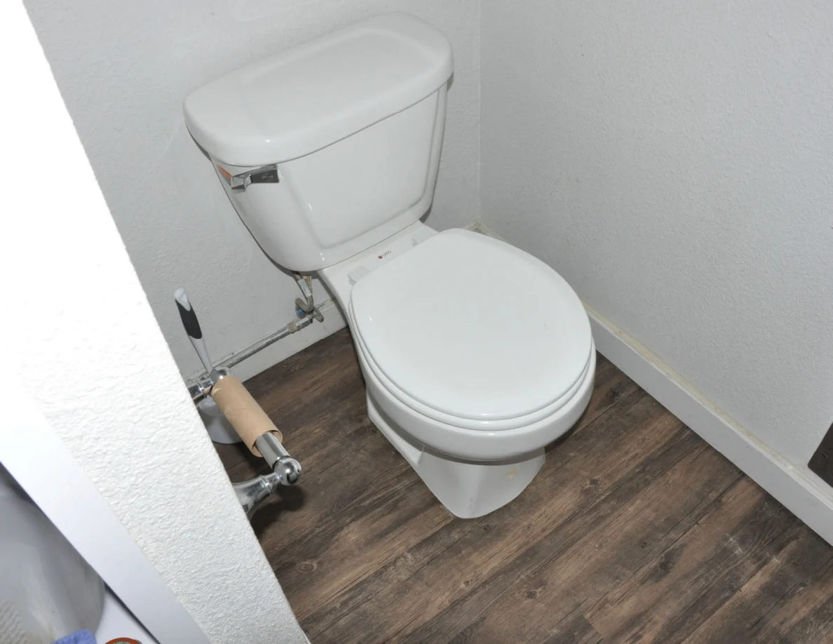 White toilet with closed lid and empty toilet-paper holder on wood-look flooring.