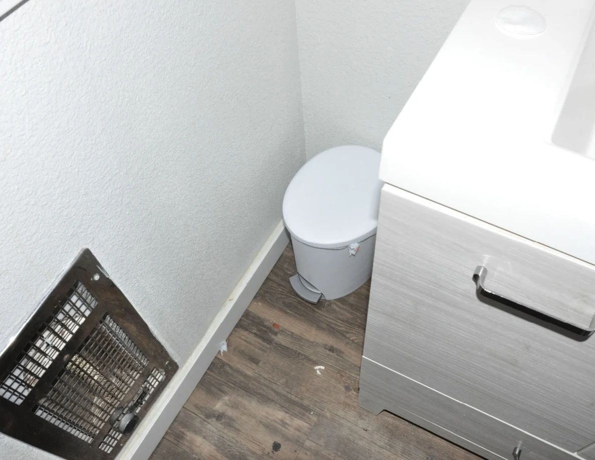 Small lidded trash bin between a bathroom vanity and wall heater.