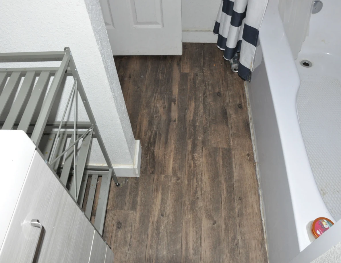 Bathroom floor beside a tub with striped curtain and narrow metal shelf.