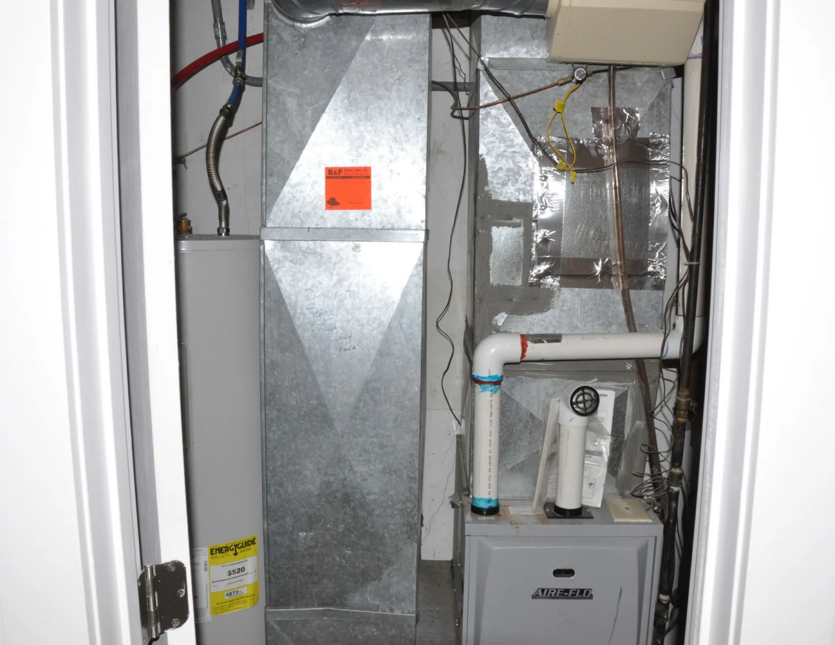 Utility closet containing a furnace, metal ductwork, pipes and exposed wiring.