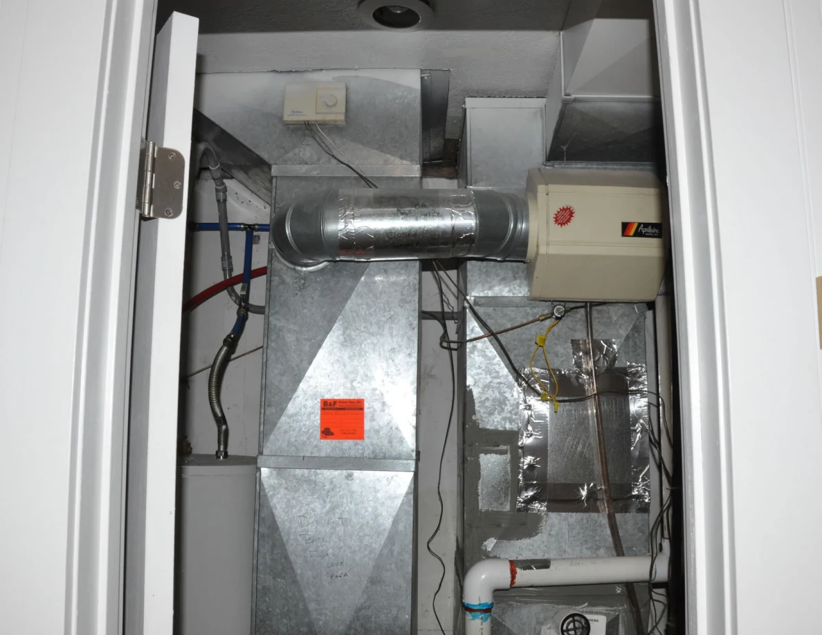 Upper utility closet with galvanized ducts, a ventilation unit and exposed pipes.