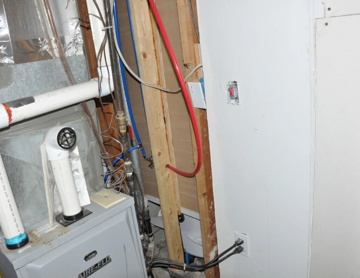 Utility-room framing with red and blue plumbing lines, wires and white pipes above a furnace.