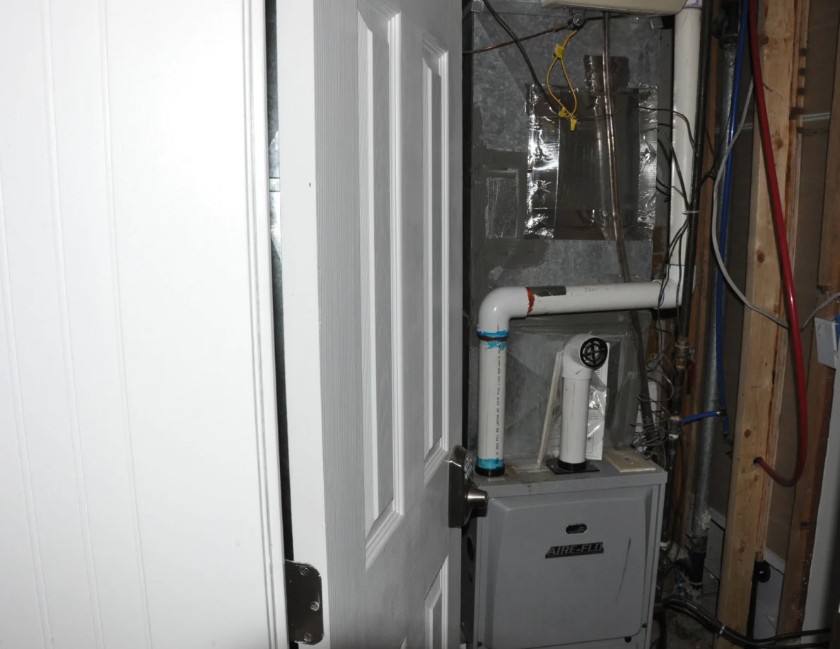 Partly open utility door with furnace, white pipes and wiring visible.