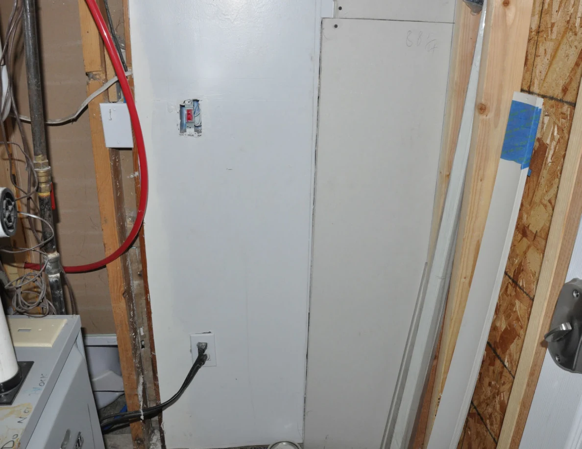 Exposed framing and red pipe beside a white panel, electrical switch and plugged-in cables.