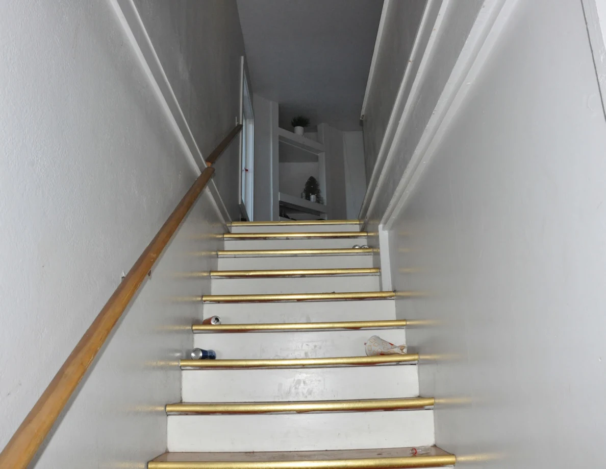 Upward view of a narrow staircase with a handrail and shelving at the top.