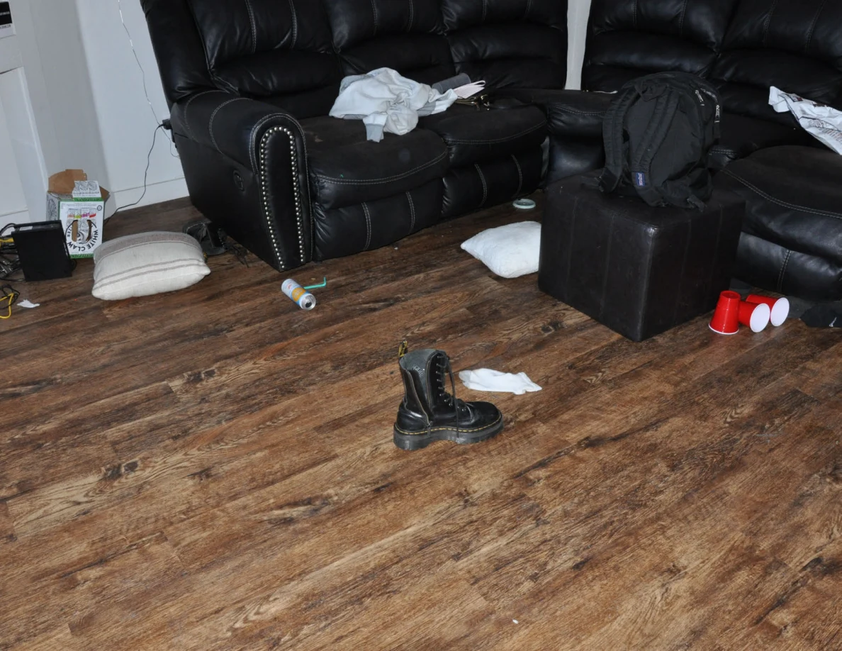 Black sofas and ottoman with a backpack, cushions, boots and red cups scattered across the wood-look floor.