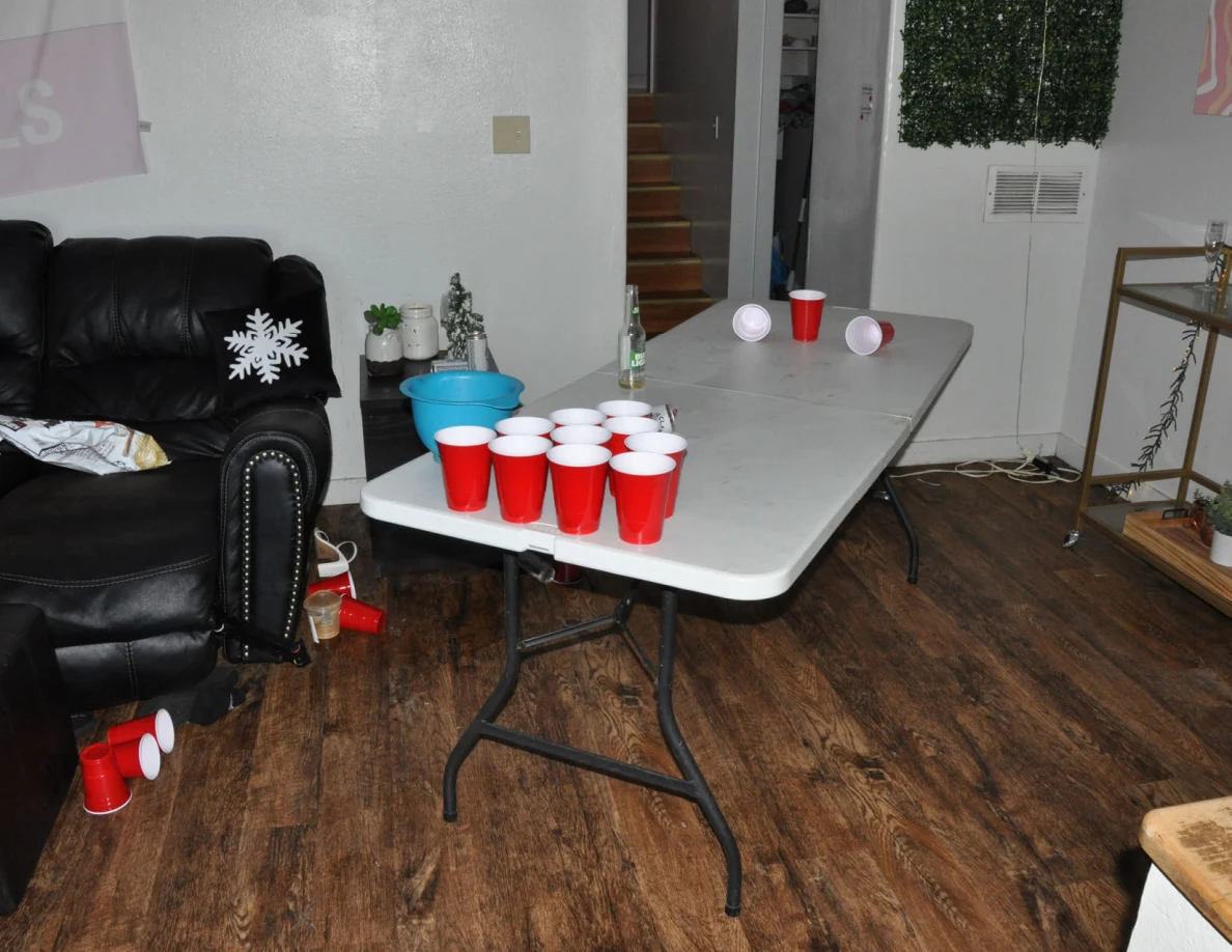 White folding table set up for cup pong with clustered red cups, a blue bowl and bottle beside a black sofa.