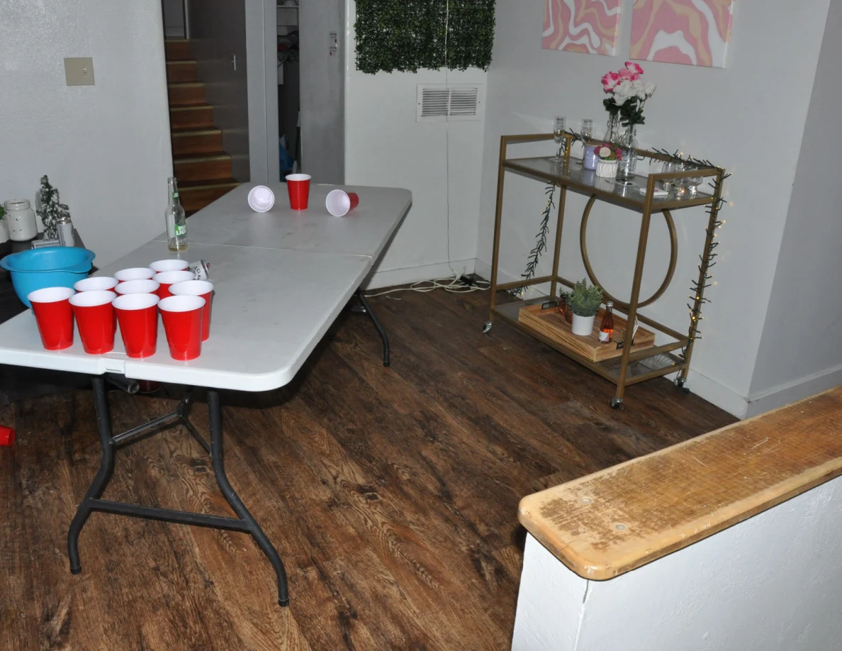 Cup-pong table with red cups beside a gold-colored cart holding flowers, bottles and a small plant.