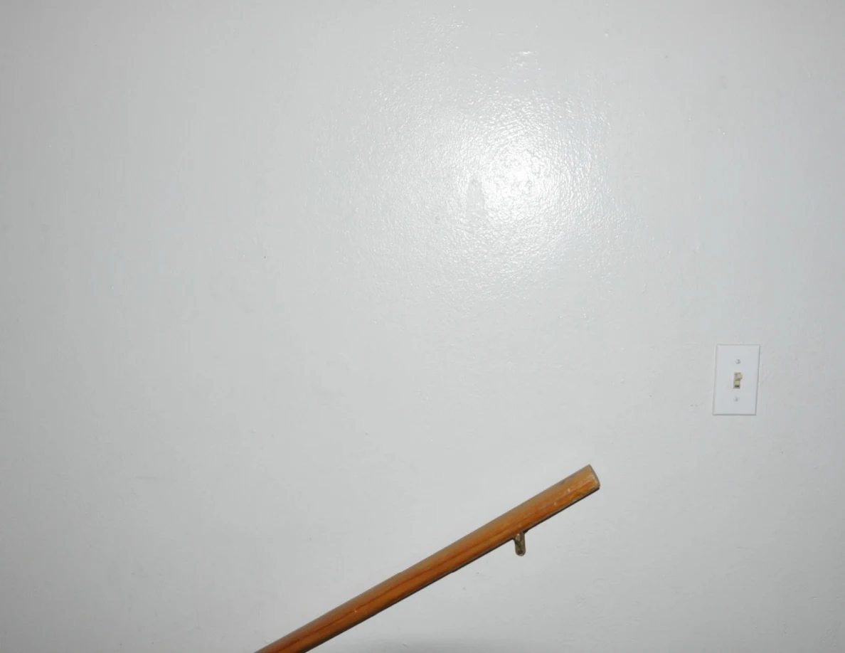 White wall and light switch above the end of a wooden stair handrail.
