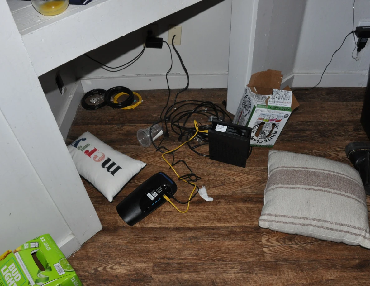 Cushions, tangled cables, electronic devices and a drink carton on the floor beneath shelving.