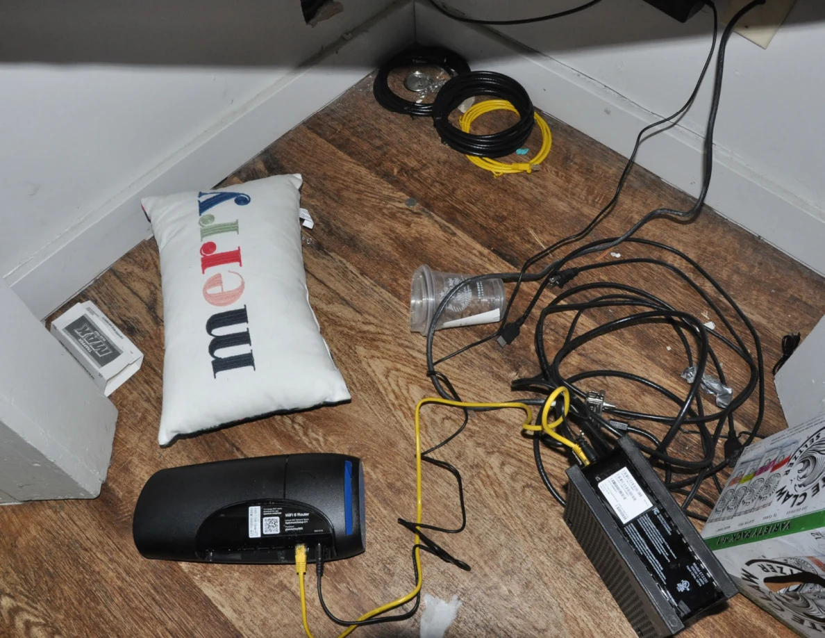 Tangled cables and electronic devices on the floor beside a Merry cushion and clear cup.
