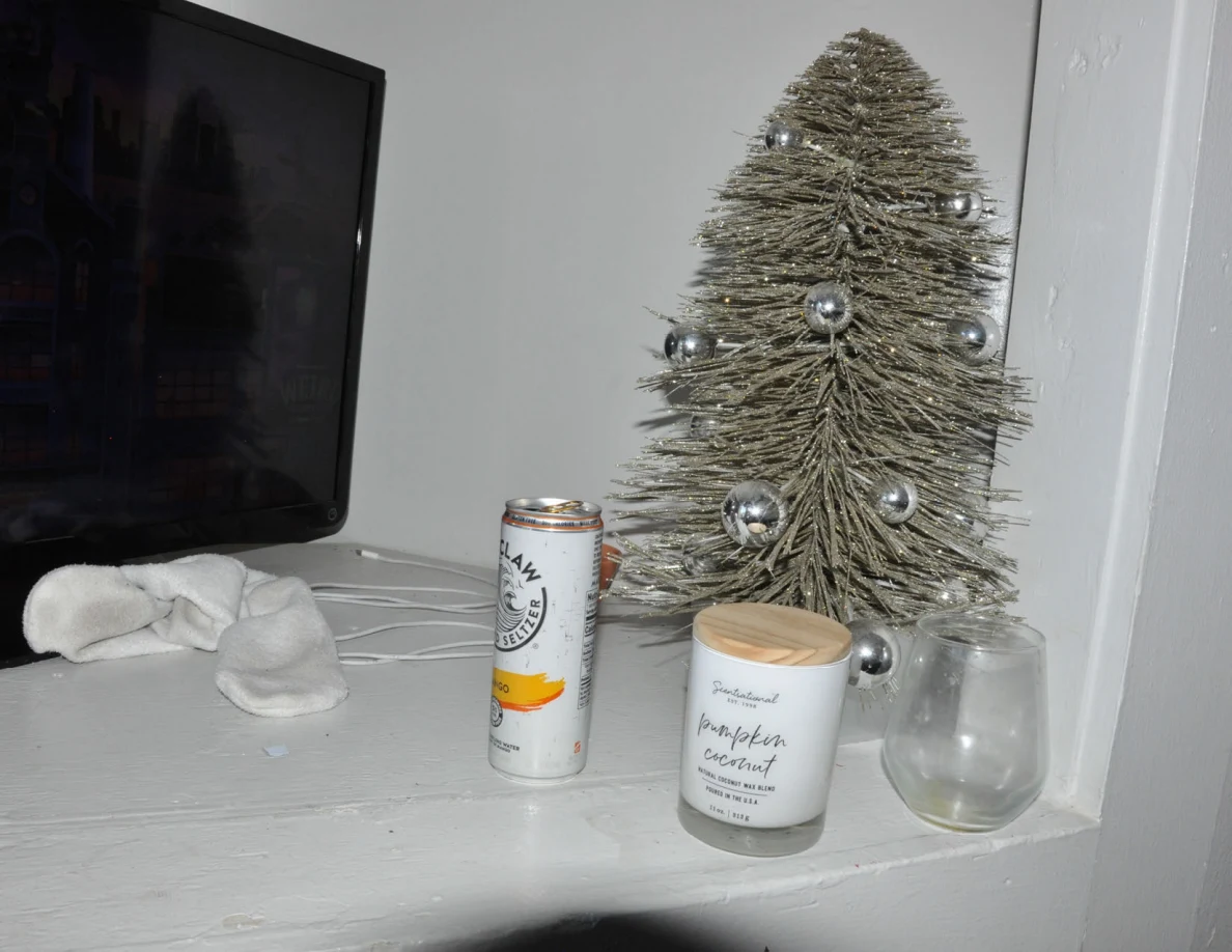 Small decorated tabletop tree beside a candle, glass, drink can and television.
