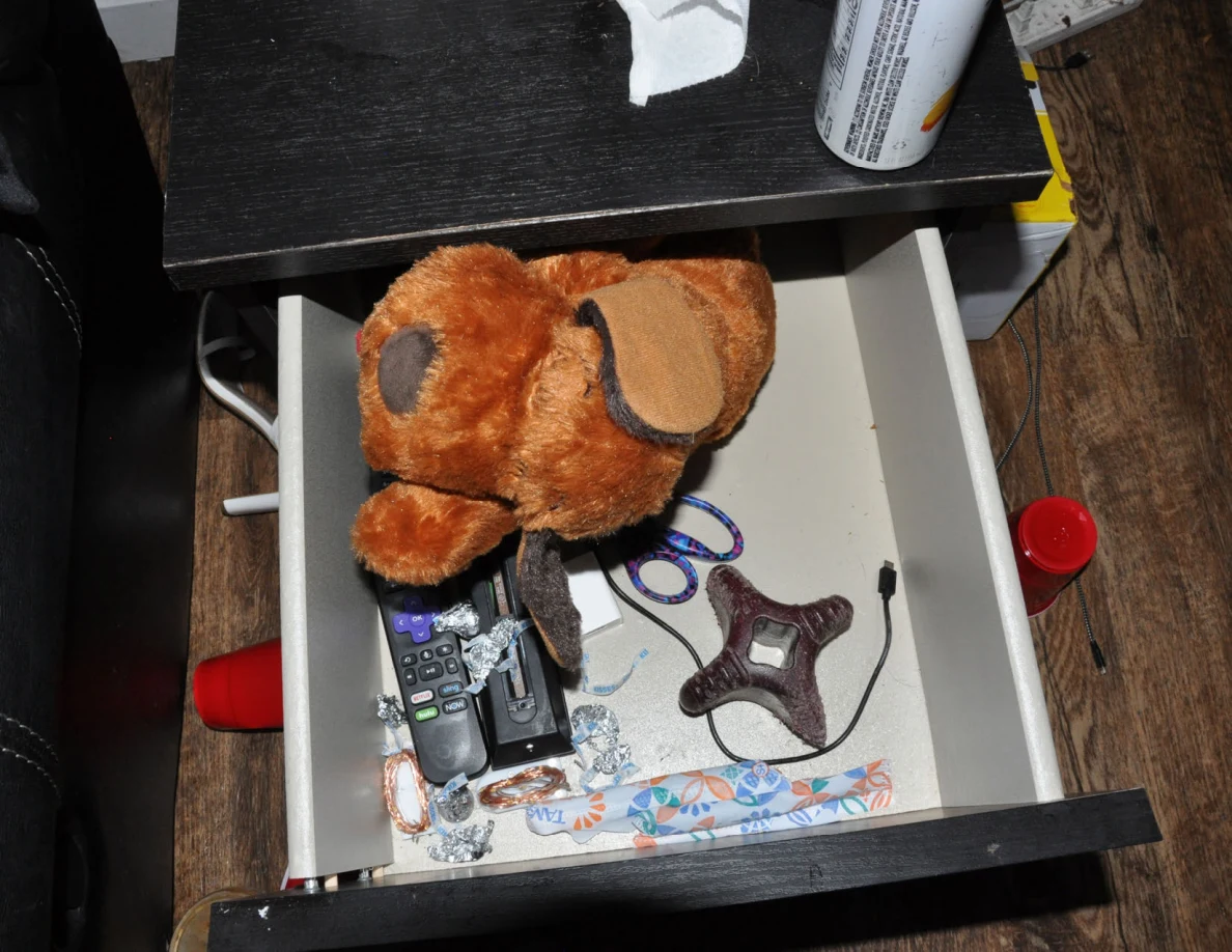Open drawer containing a stuffed dog, scissors, remote controls, cables and small objects.
