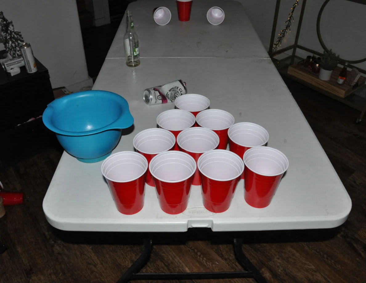 Cup-pong table with a triangular arrangement of red cups, a blue bowl, drink can and bottle.