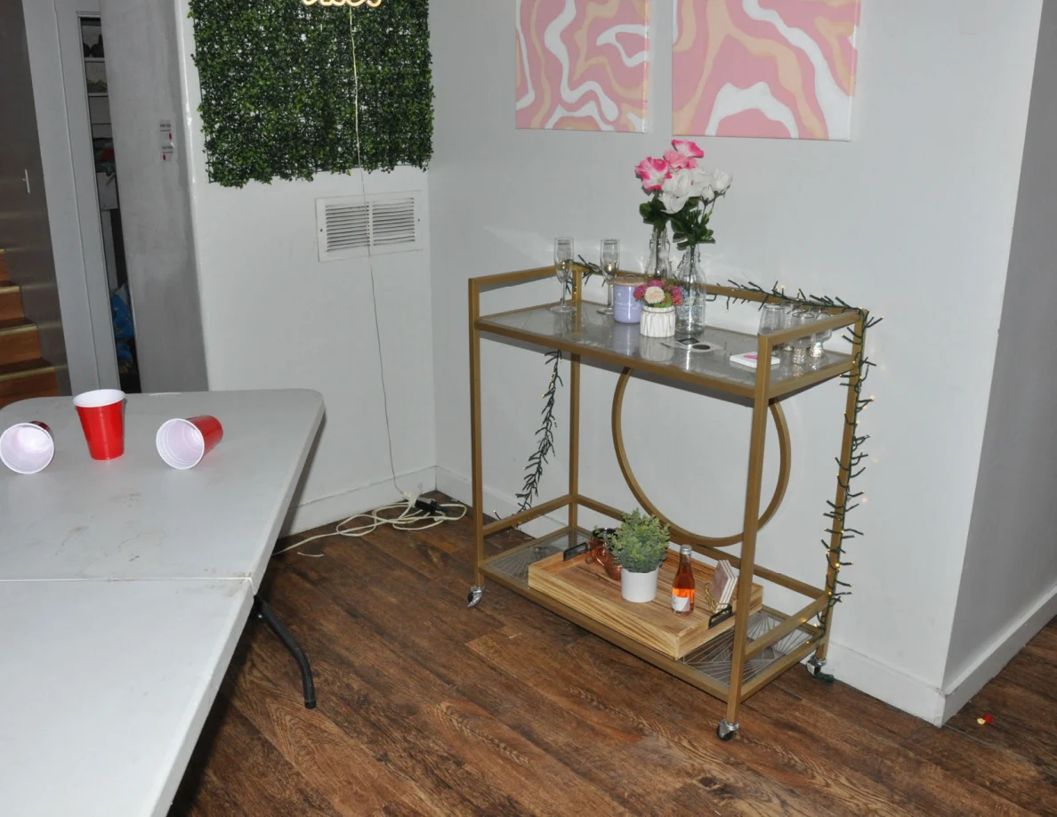 Gold-colored drinks cart holding flowers, glassware and plants beside the cup-pong table.