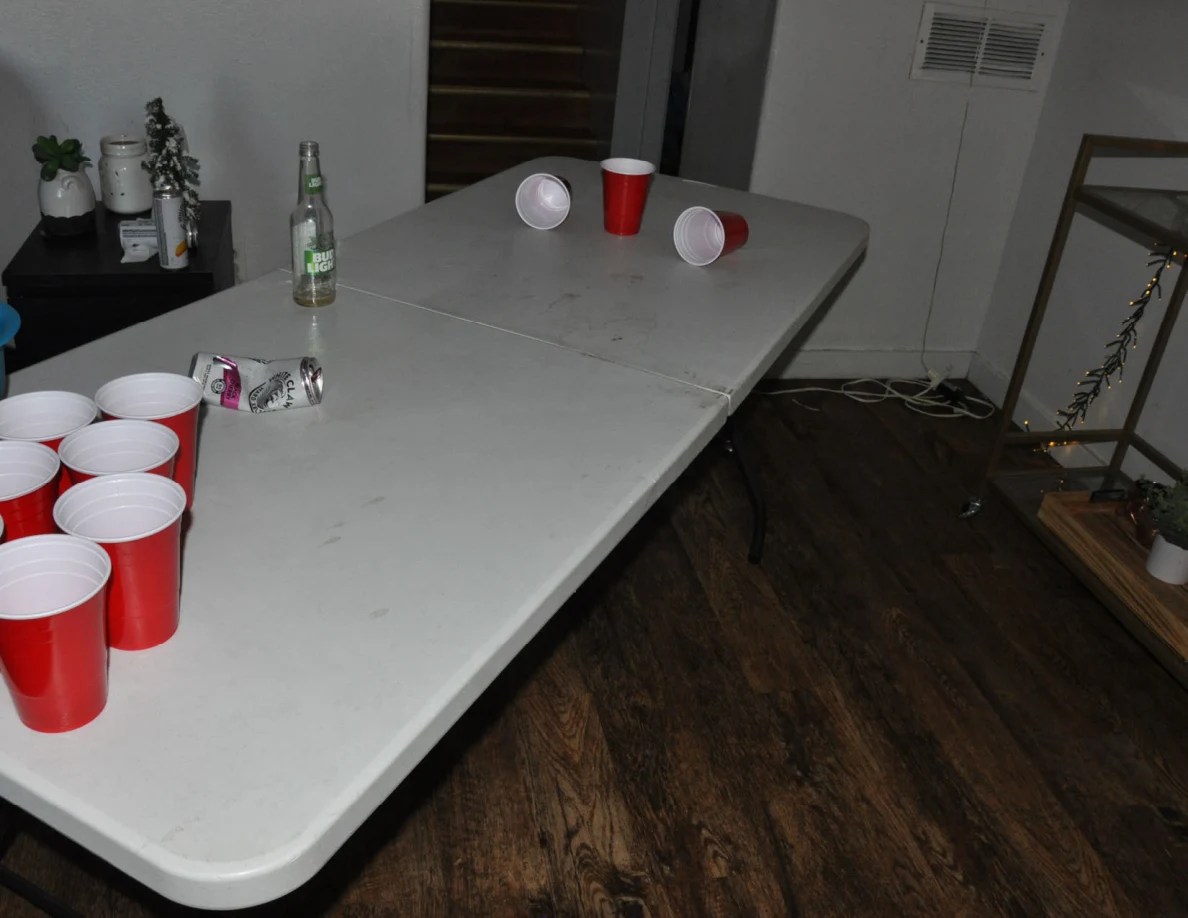 Angled view of a white cup-pong table with red cups, a bottle and crushed can.