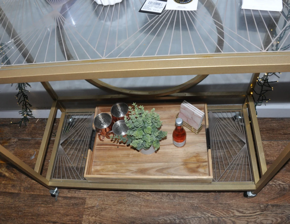 Lower shelf of a drinks cart with a plant, metal cups, small bottle and napkins on a wooden tray.