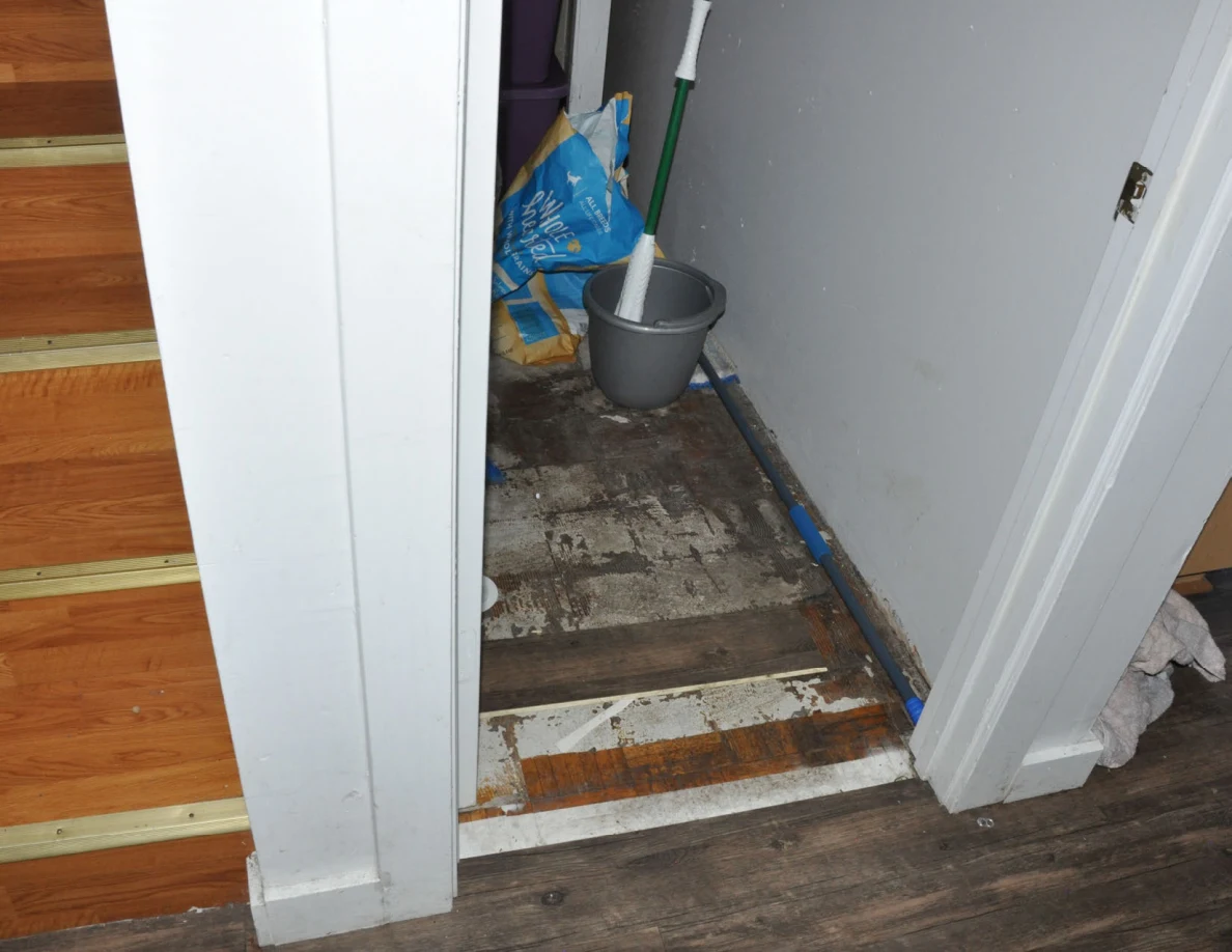 Open storage closet beside stairs with a mop, bucket and bags on a worn floor.
