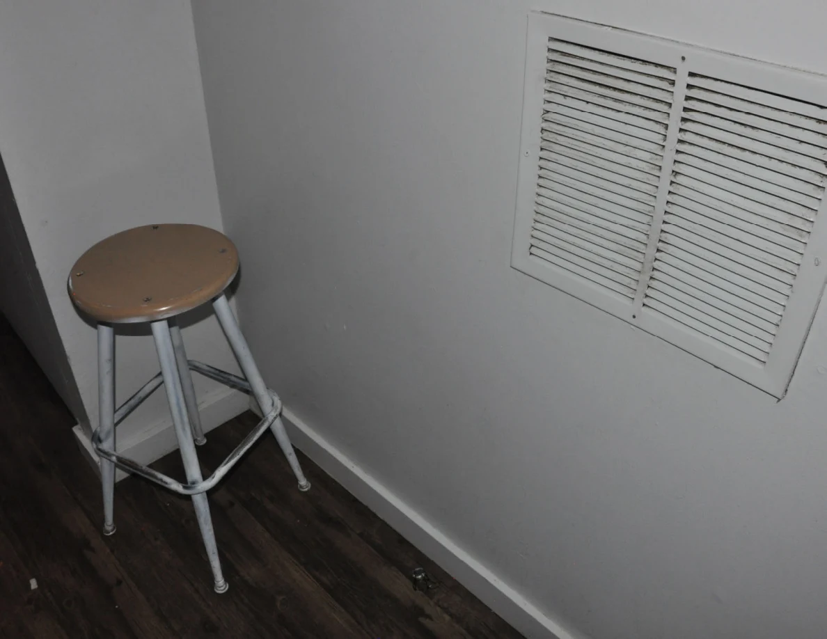 Tall wooden-seat stool beside a white wall ventilation grille.
