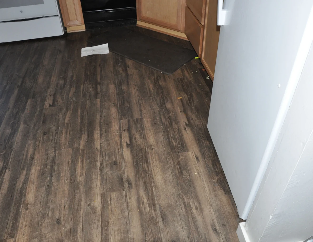Wood-look kitchen floor beside refrigerator and cabinets, with a dark mat and paper.