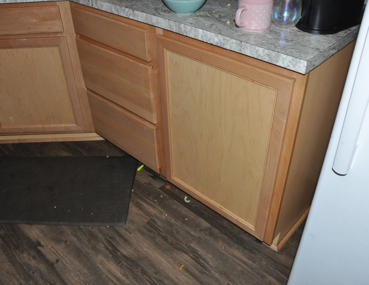 Lower kitchen cabinets with a dark floor mat and small debris near the base.