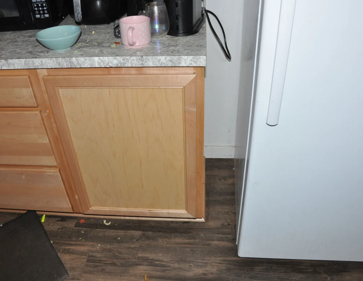 Wooden kitchen cabinet below a counter with bowl and mug beside the refrigerator.