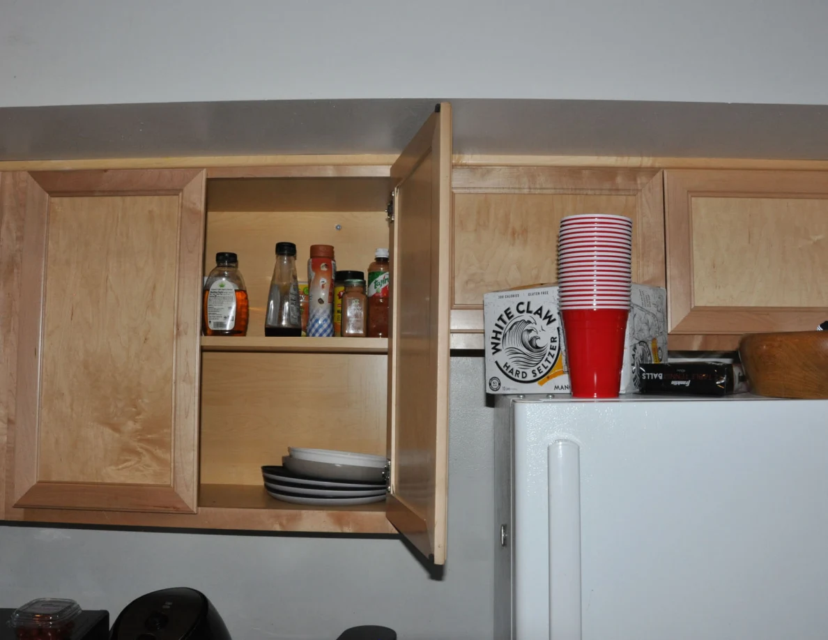 Open upper kitchen cabinet holding condiment bottles and plates beside stacked cups on a refrigerator.