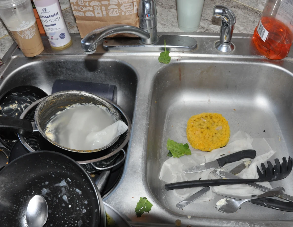 Double kitchen sink filled with dishes, pots, utensils, sponge and food scraps.