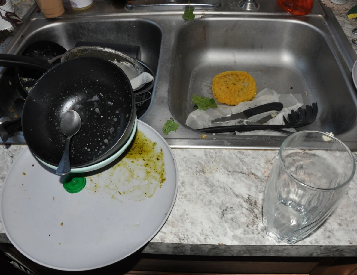 Dishes piled around a kitchen sink with a yellow sponge, utensils and leafy food scraps.