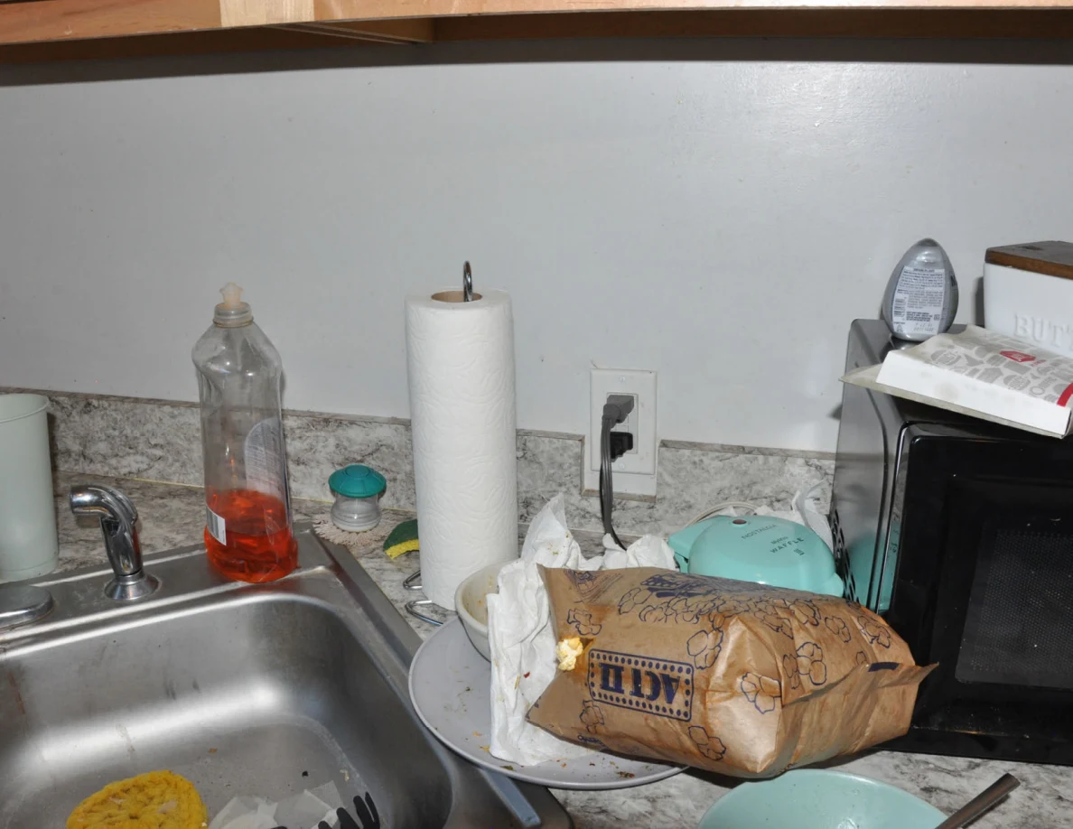 Kitchen counter beside sink holding dish soap, paper towels, paper bag and dishes.