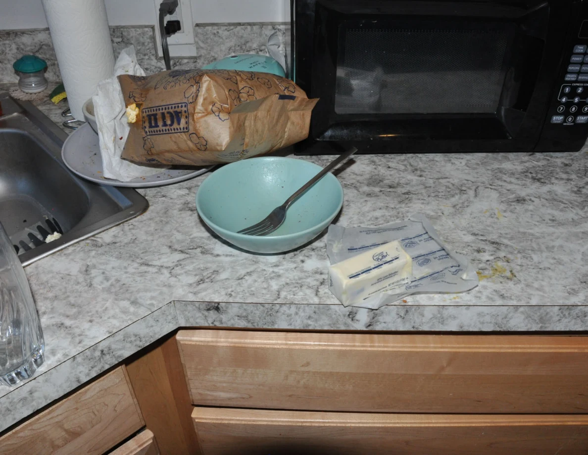 Kitchen counter with turquoise bowl and fork, wrapped butter, paper bag and black microwave.