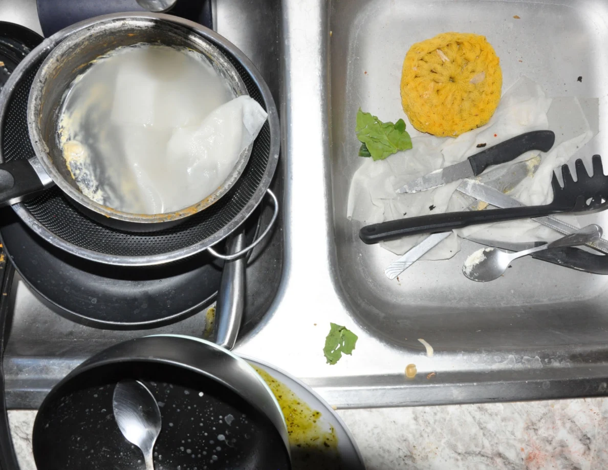 Close view of sink containing pots, utensils, sponge and leafy food scraps.