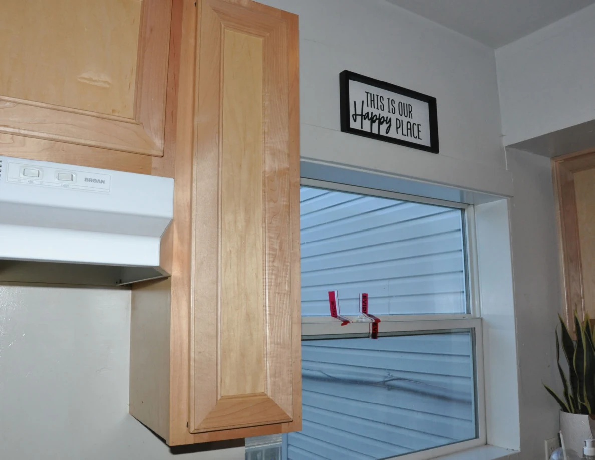 Tall closed wooden cabinet beside a kitchen window and This Is Our Happy Place sign.
