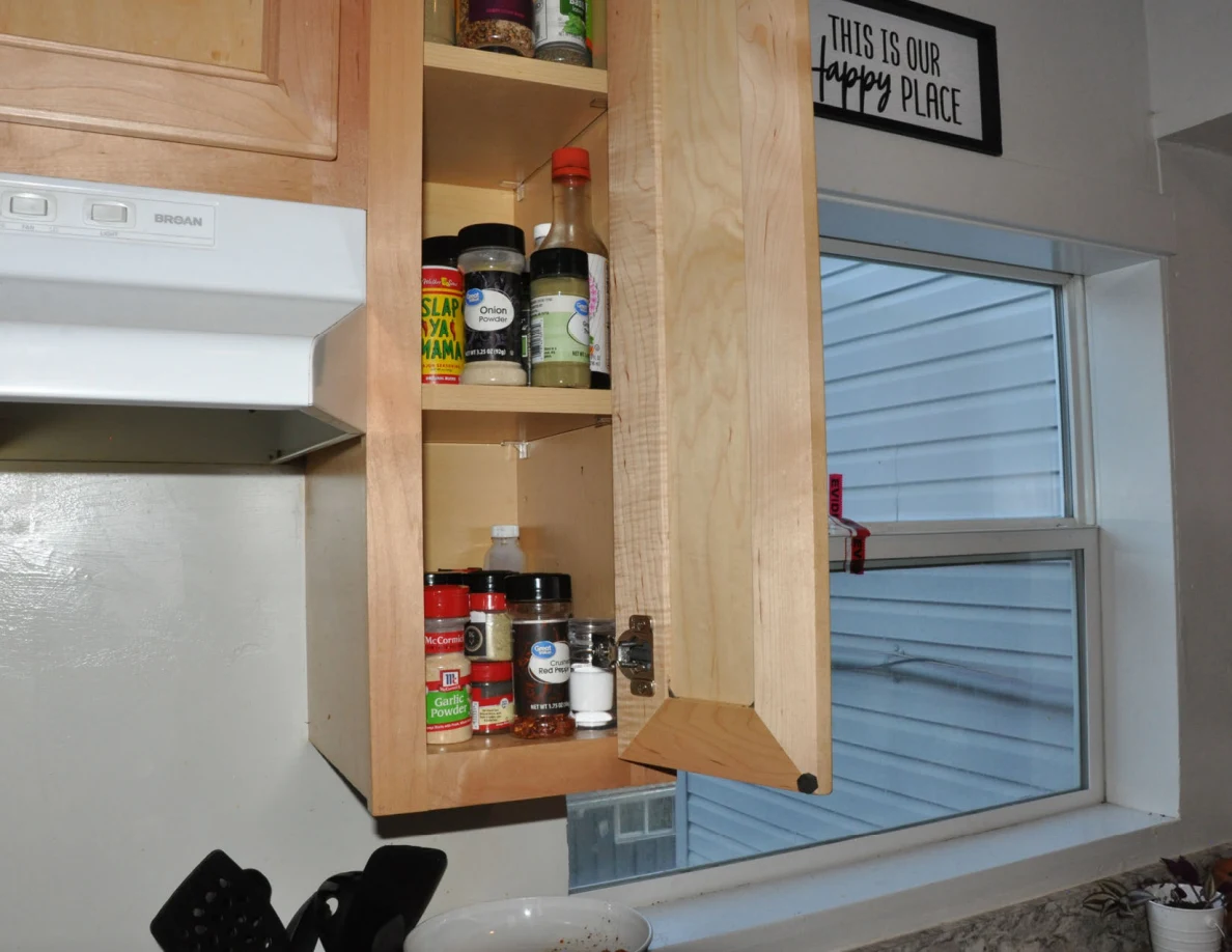 Open narrow cabinet filled with spice jars and bottles beside a kitchen window.